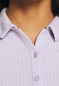 Camisa de color lila con relieve que presenta un cuello con solapa y un cierre de botones con botones redondos y translúcidos. Textura sutil visible en la tela.