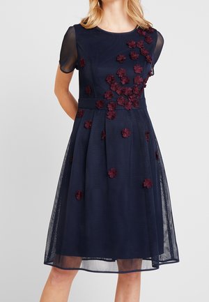 Kobieta ubrana w granatową sukienkę do kolan z krótkimi przezroczystymi rękawami i bordowymi kwiatowymi aplikacjami rozmieszczonymi na przodzie i w talii.