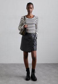 Top de manga larga a rayas en negro y crema, combinado con una falda envolvente de cuero negro. La modelo lleva botas negras con cordones y sostiene un pequeño bolso de color oliva.