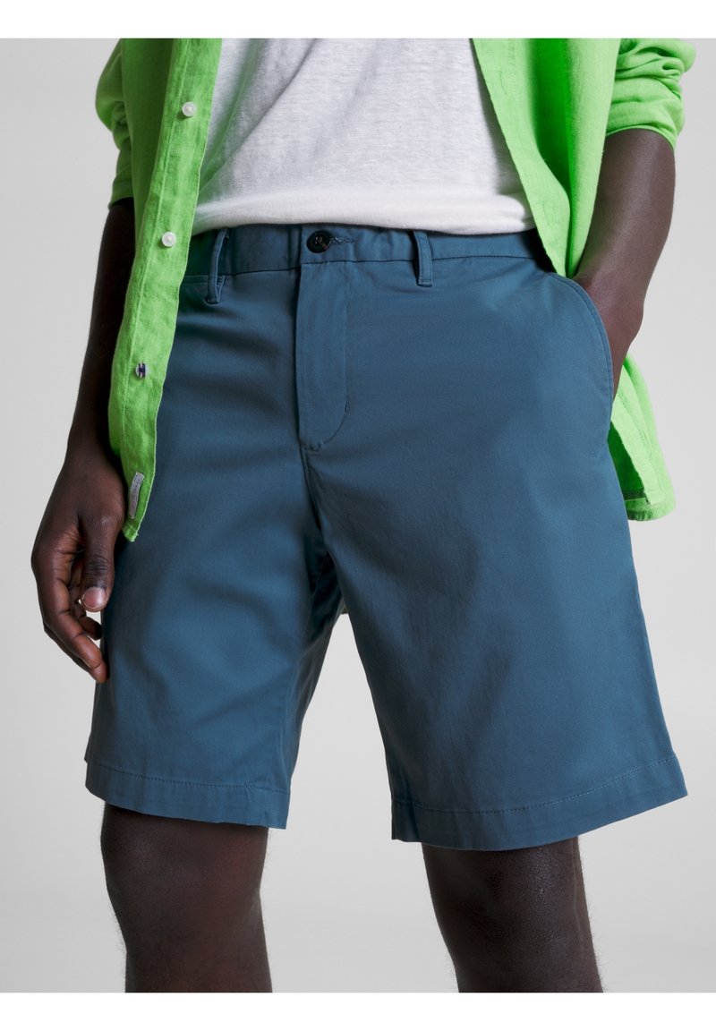 Homme portant un short bleu, un t-shirt blanc et une chemise verte déboutonnée, une main dans une poche sur un fond uni.