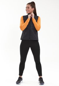 Schwarze ärmellose Weste mit puffiger Textur, Reißverschluss vorne und seitlichen Taschen, getragen über einem orangefarbenen Langarmoberteil und schwarzen Leggings.