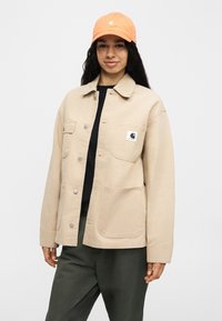 Νεαρό άτομο που φοράει μπεζ σακάκι με κουμπιά και εμπρόσθιες τσέπες, κονκάρδα με λογότυπο, μαύρο πουκάμισο, σκούρο παντελόνι και πορτοκαλί καπέλο μπέιζμπολ.