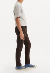 Maglietta grigia in cotone, pantaloni marroni a taglio slim e sneakers in tela blu con suole bianche. Nessun motivo o dettaglio visibile.