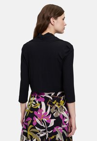 Model met lang bruin haar draagt een zwarte top met een gerimpelde achterkant en een bloemenrok met paarse en groene patronen.