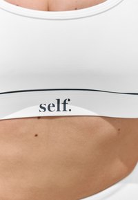 Bijeli sportski grudnjak s širokim pojasom na kojem je crni tekst "self." Tkanina izgleda glatko s prilagođenim dizajnom.