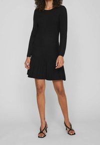 Donna in un abito lungo a maniche lunghe nero a coste con gonna plissettata, che indossa sandali con tacco neri a fasce, in piedi su uno sfondo grigio uniforme.