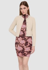 Cardigan cropped beige con una texture morbida, caratterizzato da un collo rotondo e chiusura con bottoni. Abbinato a un vestito marrone fitted con motivo floreale rosa.