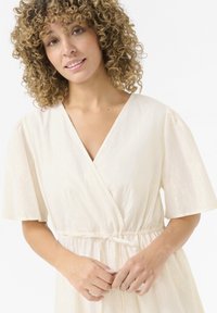 Light beige, knee-length wrap dress with short puff sleeves, a v-neck, and a cinched waist. Textured fabric with a subtle shimmer.