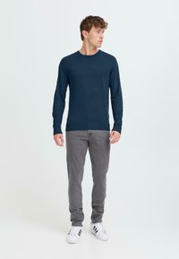 Námornícky modrý sveter s dlhými rukávmi z jemného materiálu, s rebrovaným lemom a manžetami, v kombinácii so svetlošedými slim-fit džínsami a bielymi teniskami s čiernymi pruhmi.