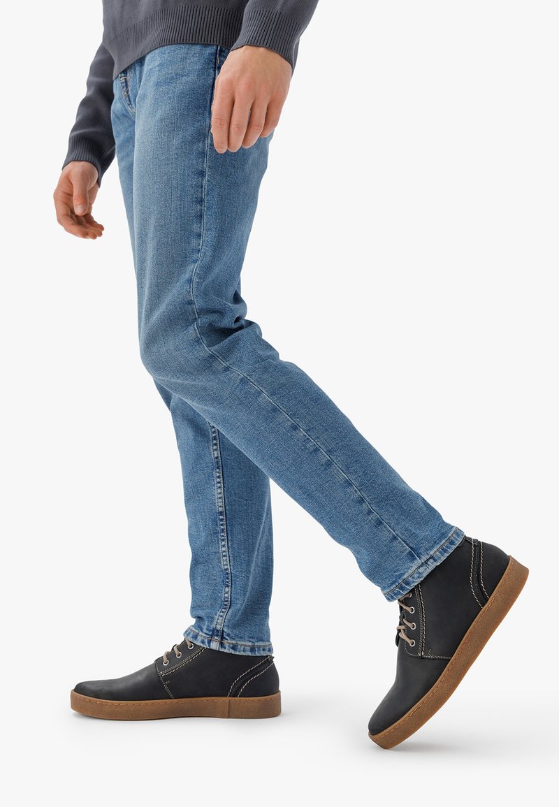 Scarpe in pelle nera con suole in gomma marrone, abbinate a lacci grigi, indossate con jeans in denim blu, enfatizzando un look informale.