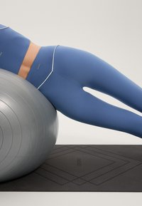 Modra aktivna majica in legice z belimi detajli, ležijo na sivi fitnes žogi na teksturirani črni podlagi z geometrijskim vzorcem.