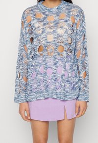 Lyseblå strikket sweater med store åbne huller og blomsterdesign, parret med en lys lilla nederdel med slids i siden.
