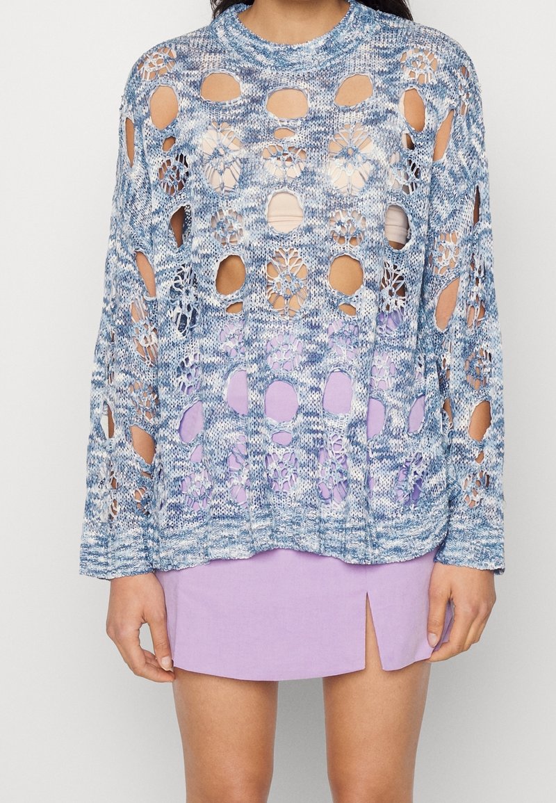 Lyseblå strikket sweater med store åbne huller og blomsterdesign, parret med en lys lilla nederdel med slids i siden.