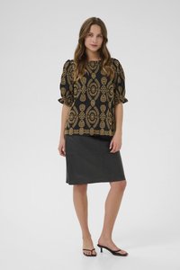 Svart blus med guld geometriskt mönster, puffärmar; i kombination med en figurnära svart läderkjol. Modell bär svarta klacksandaler.