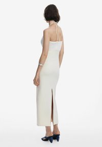 Long cream dress with a fitted bodice, halter straps, and a side slit. Worn with blue heeled sandals and a silver bracelet on the wrist.