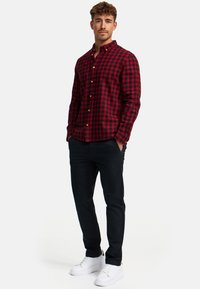 Camisa de cuadros rojos y negros con cuello abotonado, combinada con pantalones oscuros y zapatillas blancas, exhibida en un modelo sobre un fondo blanco.