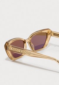 Gennomsiktlig amber katteøyne solbriller med teksturerte armer og lilla tonede linser som hviler på en hvit overflate.