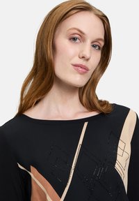 Schwarze Tunika mit geometrischen Mustern in Beige und Braun. Verziert mit Strasssteinen, hat sie lange Ärmel und einen weiten Ausschnitt.