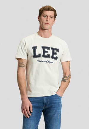 Mężczyzna z krótkimi włosami i tatuażami ma na sobie białą koszulkę "LEE American Original" i niebieskie dżinsy, stojąc z jedną ręką w kieszeni.