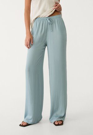 Kvinde iført lyseblå viddebukser med elastik og snøre i taljen, kombineret med en cremefarvet top og sorte sandaler med åbne tæer.