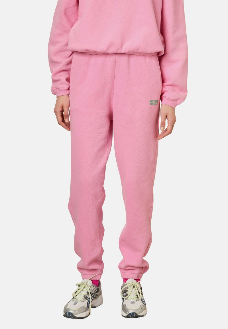 Pantalon de survêtement rose en tissu polaire doux avec une taille élastique et des poignets. Présente un petit logo vert sur la hanche gauche.