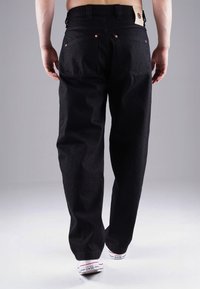 Schwarze Jeans mit geradem Bein und zwei Gesäßtaschen. Mit Kupferriegeln und einem Lederpatch am Bund. Getragen mit weißen Sneakern.