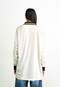 Krémová bunda s geometrickým diamantovým vzorem, černým límcem a žlutými detaily. Dlouhé rukávy s černými manžetami.