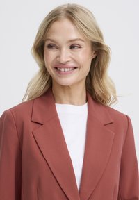 Smiling blonde woman looking to the side, wearing a rust-colored blazer over a white top, against a plain light background.