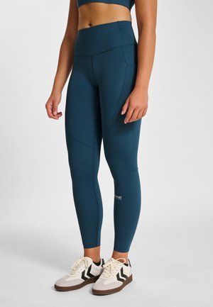 Højttaljede leggings i teal med tætsiddende design, sidesømme og logodetalje. Bærtes med hvide og sorte sportssko på en neutral baggrund.