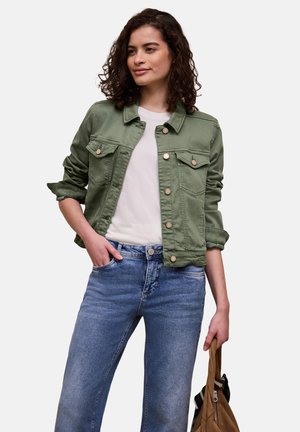 Kvinde iført grøn jakke med knapper, hvid skjorte og blå jeans, med en brun taske i venstre hånd.