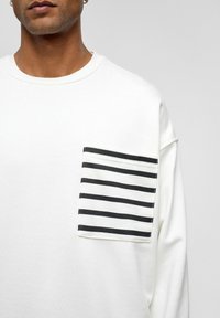 Witte sweatshirt met een zwart-wit gestreepte zak. Zacht katoenmateriaal, ronde halslijn, ontspannen pasvorm en zichtbare stikselaccenten.