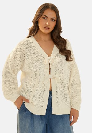 Crème gebreide cardigan met een open voorkant met twee strikjes, een ruitpatroon en ballonmouwen. De textuur is lichtgewicht en luchtig.