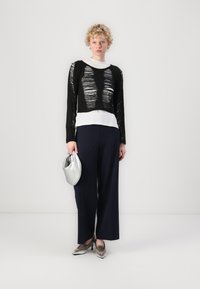 Schwarzes Langarmoberteil mit horizontalen Ausschnitten, weißer Unterlage, marineblaue weit geschnittene Hose, metallische silberne Tasche und silberne Schuhe.