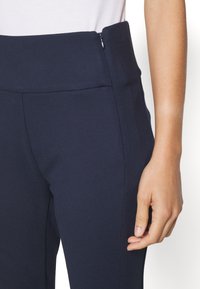 Pantalons bleu marine avec une taille haute, dotés d'une fermeture éclair sur le côté et d'une coupe structurée, fabriqués dans un tissu lisse et texturé.