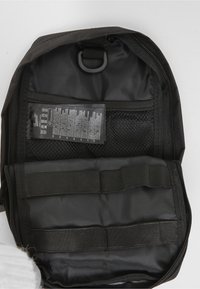 Bolsa negra con cremallera abierta para mostrar el interior con bolsillo de malla, correas elásticas y etiqueta de cuidado de la tela fijada en la tapa interior.