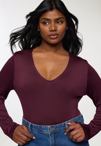 Bordowy body z długimi rękawami i głębokim dekoltem w kształcie V, zestawione z niebieskimi dżinsami z wysokim stanem. Gładki materiał, codzienny fason.