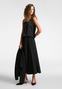 Top in satin nero abbinato a una gonna nera fluente. Gli accessori includono un blazer nero e scarpe con tacco marroni a punta.