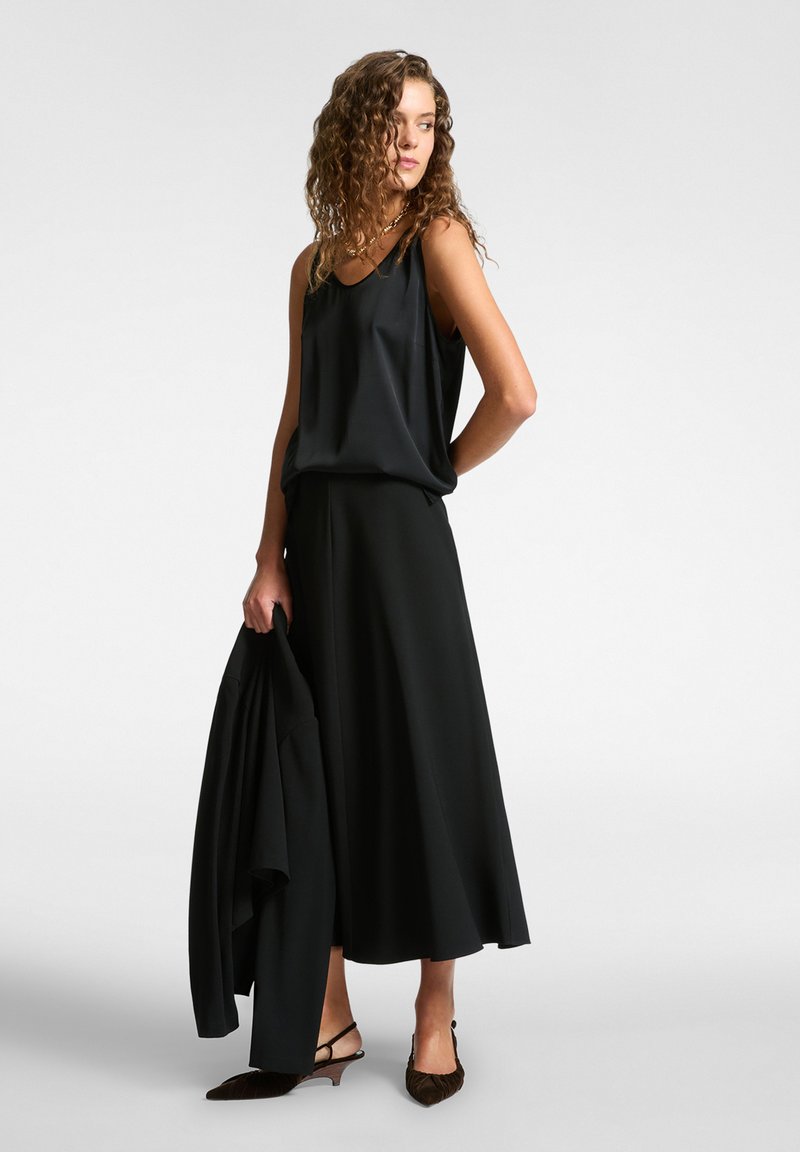 Top in satin nero abbinato a una gonna nera fluente. Gli accessori includono un blazer nero e scarpe con tacco marroni a punta.