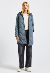 Ljusblå quiltad jacka med huva, med en framsidans dragkedja och tryckknappar; bärs över en krämfärgad tröja och svarta byxor.