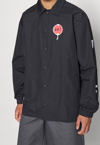 Svart coach-jacka med knäppning, elastiska ärmslut och en cirkulär röd logotyp på vänster bröst samt vita detaljer på sidorna.