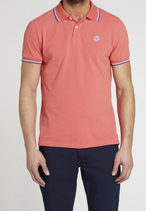 Mężczyzna w koralowej koszulce polo z białym i granatowym wykończeniem na kołnierzyku i rękawach, w ciemnogranatowych spodniach, stojący z rozluźnionymi rękami.