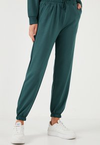 Pantaloni della tuta teal con vita elastica, polsini arricciati e tasche laterali, abbinati a sneaker bianche. Tessuto morbido e leggero.