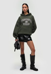 Felpa verde con testo bianco, mini gonna nera con texture lucida, stivali neri alti fino al ginocchio con dettagli in argento, che tiene una piccola borsa nera.