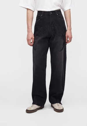 Personne portant un jean noir à jambes larges avec un motif embossé subtil, associé à des baskets blanches et beiges, debout devant un fond uni.
