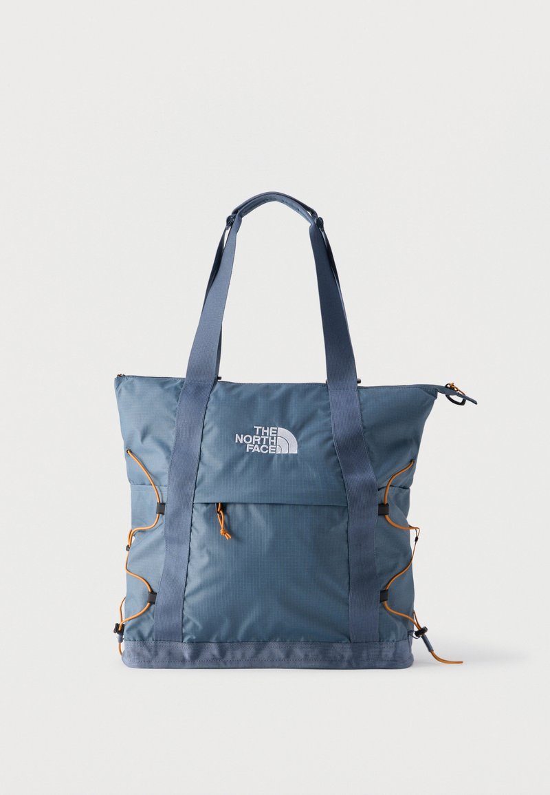 Μπλε τσάντα tote The North Face με διπλούς ιμάντες ώμου, εμπρόσθια τσέπη με φερμουάρ και πορτοκαλί ρυθμιζόμενα κορδόνια στα πλαϊνά, πάνω σε μονόχρωμο φόντο.