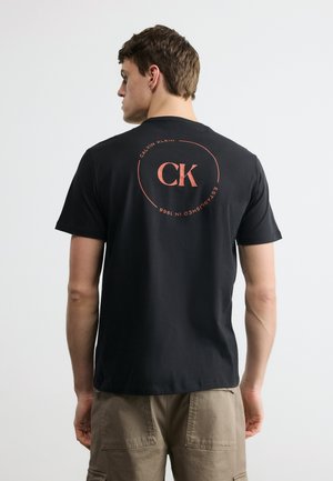 Mand iført sort Calvin Klein T-shirt med lyserødt cirkulært logo og beige shorts, vender væk mod en ensartet lys baggrund.
