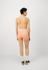 Set de brâu sportiv și pantaloni scurți de culoare piersică, realizat dintr-un material elastic; include un design tip racerback și un tiv lat de culoare albă cu logo-ul brandului.