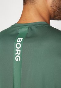 Zelené sportovní tričko s texturovanou tkaninou, kulatým výstřihem a vertikálním pruhem s bílým nápisem "BORG" na zadní straně.