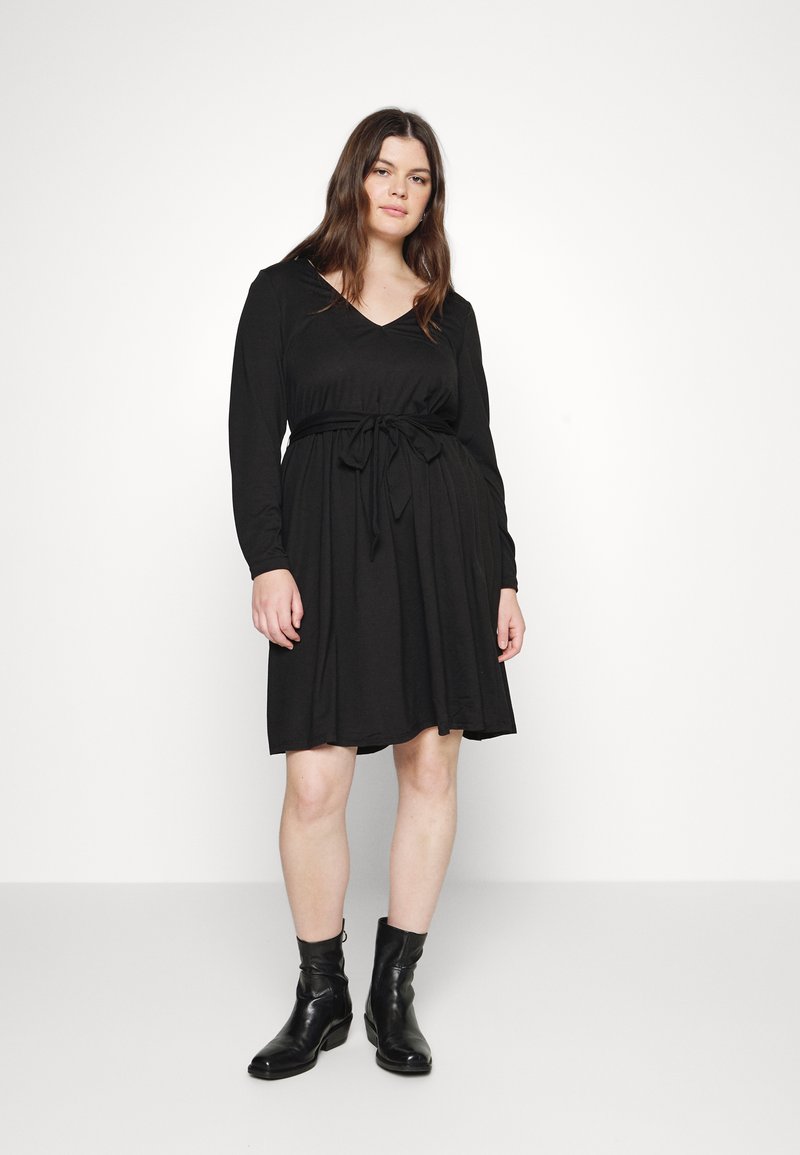 Vestido preto de mangas longas com decote em V, ajustado na cintura com um cinto de tecido combinante. Textura suave e lisa. Usado com botas pretas até o tornozelo.