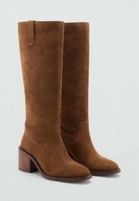 Botas de ante marrón hasta la rodilla con punta afilada, tacón apilado y una pequeña asa en la parte superior para facilitar su calzado. Textura lisa en toda su superficie.